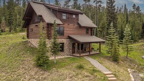 A log cabin with a stone foundation, a large porch, and a view of a forested mountain range.