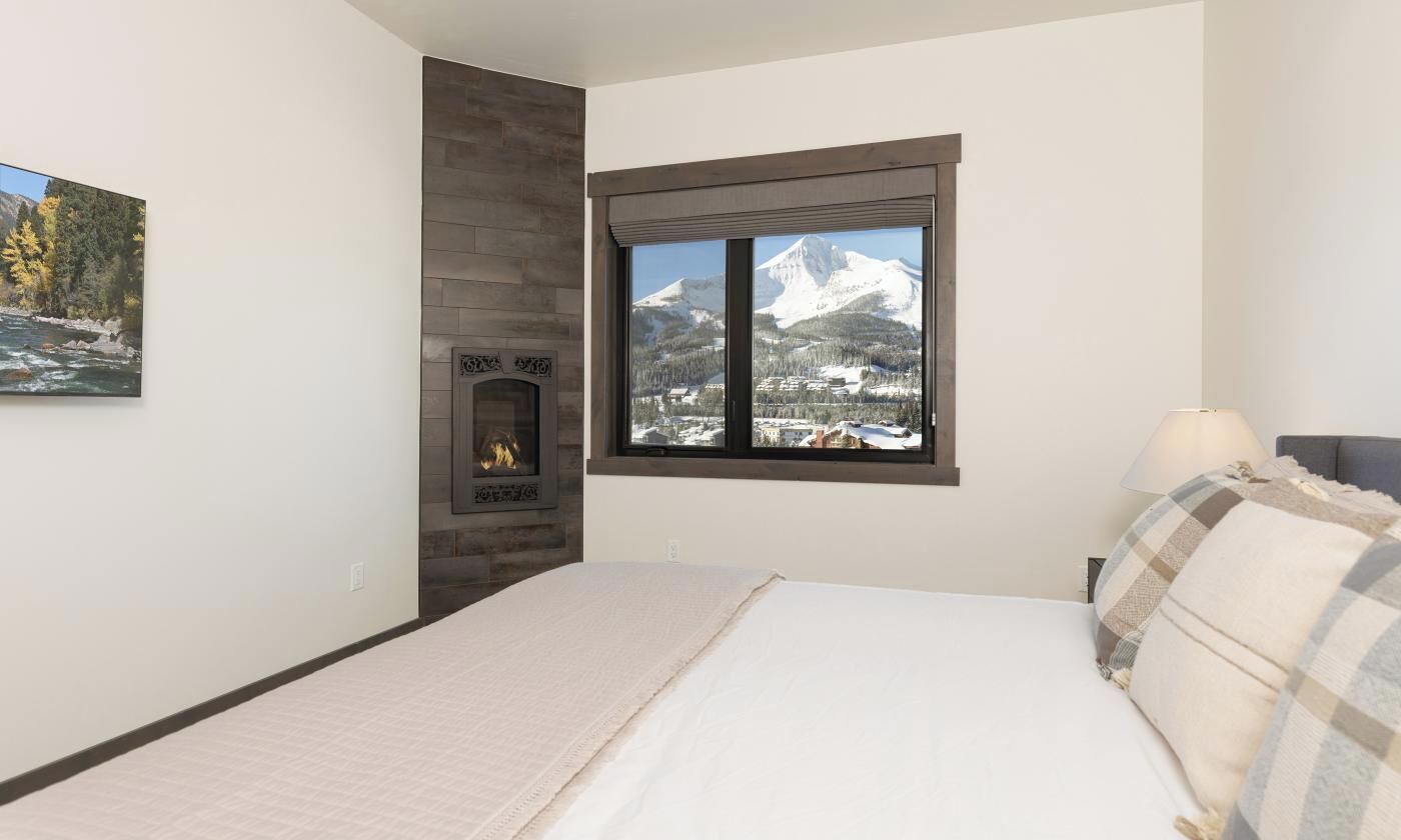 A bedroom with a fireplace, a large window with a mountain view, and a bed with a plaid blanket.