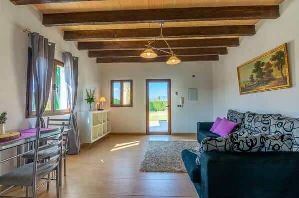 Ein Zimmer mit Holzboden, einer Couch mit gemusterten Kissen, einem Esstisch mit Stühlen und Blick durch eine Tür in einen Außenbereich.