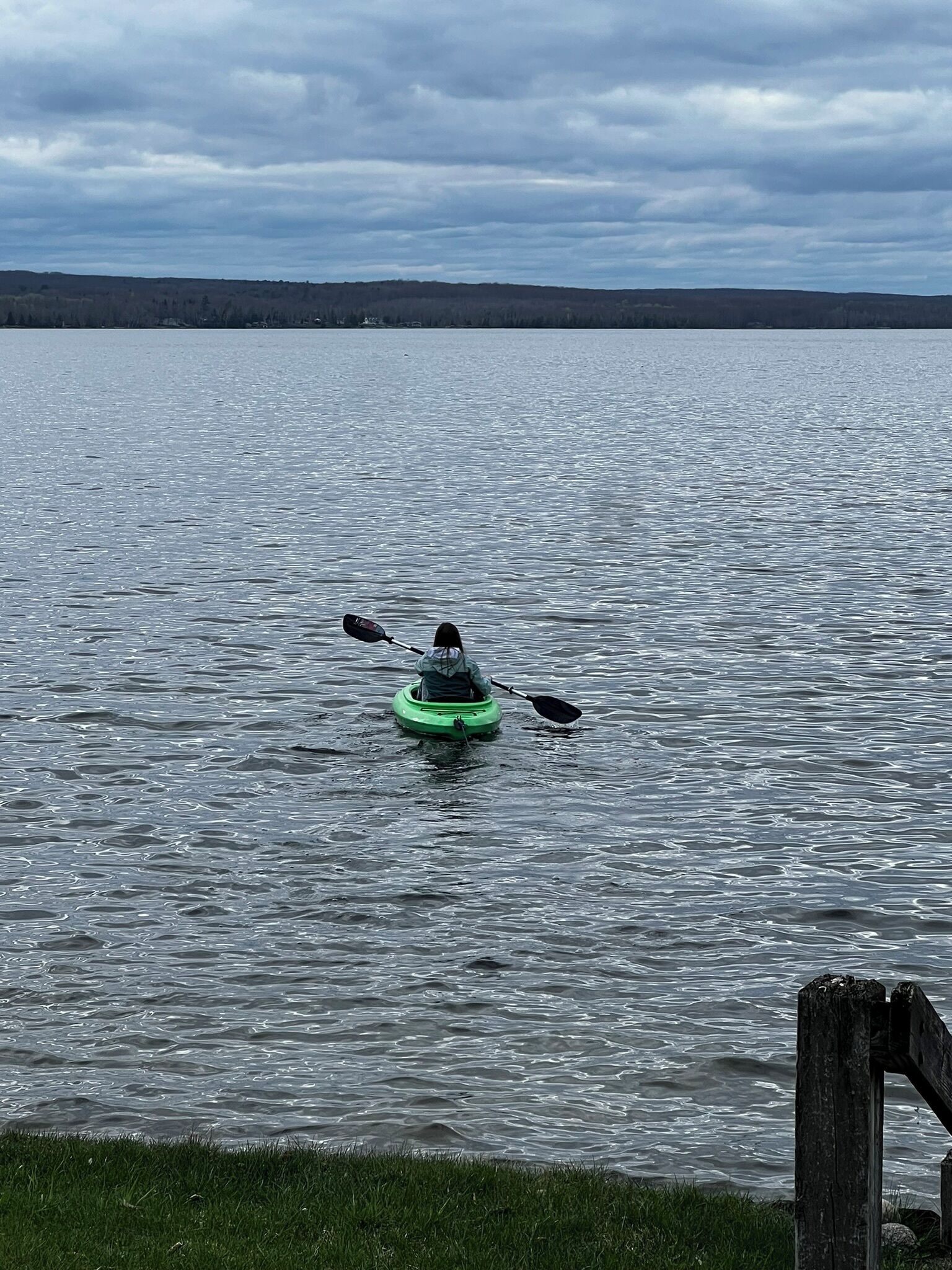 Ein Kajakfahrer in einem grünen Kajak auf einem ruhigen See mit einer grasbewachsenen Uferlinie und einem Holzsteg im Vordergrund.