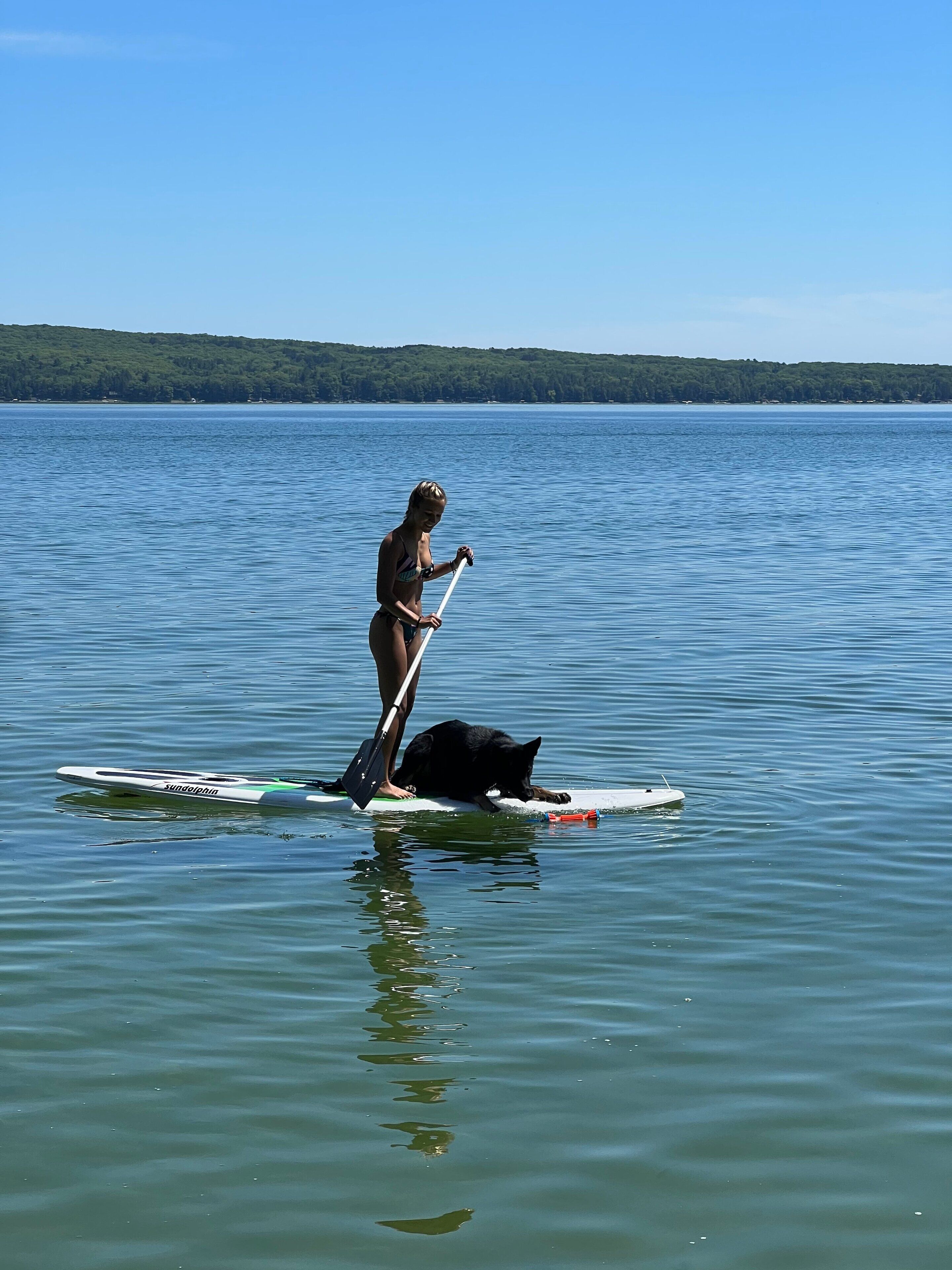 Eine Person steht auf einem Paddleboard im Wasser, während ein Hund auf dem Brett liegt.