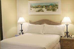 A bed with two white pillows and two bedside lamps, a wicker headboard, and a framed beach scene on the wall.