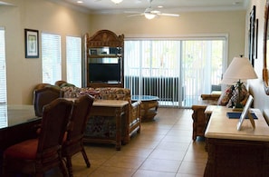A spacious living room with a television, comfortable seating, a dining area, and a large window with blinds.