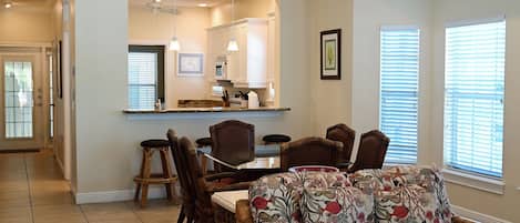 A dining area with a table and chairs, a kitchen in the background, and a window with blinds.