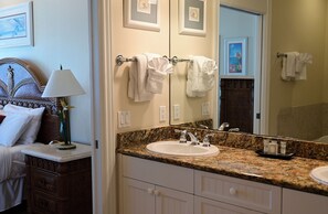 A hotel bathroom with a granite countertop sink, a mirror, and a bed with a headboard.
