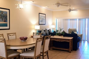 A dining area with a round table, chairs, and a decorative console table. There are ceiling fans, wall-mounted lamps, and a large window with blinds. - Spacious One-Bedroom Condo with Island Comforts (Bradenton Beach)