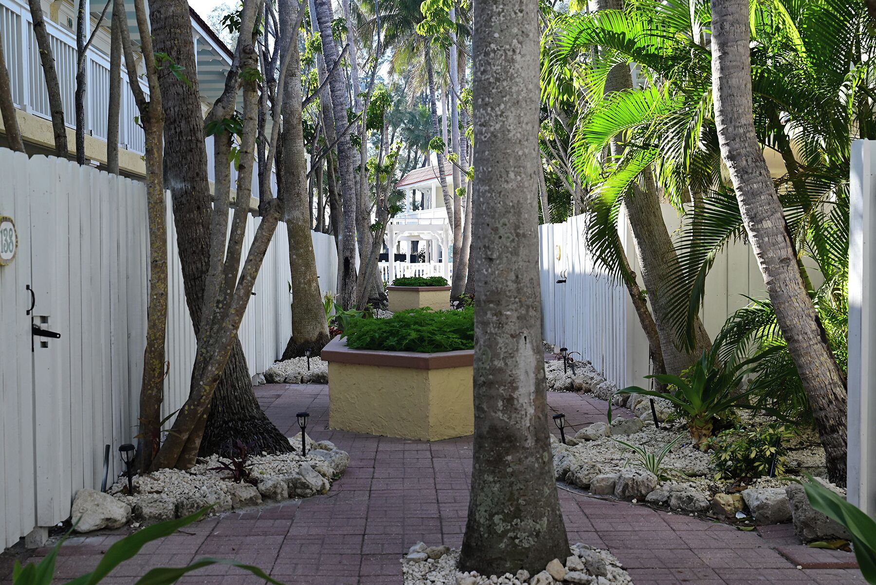 Un sendero bordeado por árboles y una cerca blanca, que conduce a un edificio con fachada blanca y un balcón.