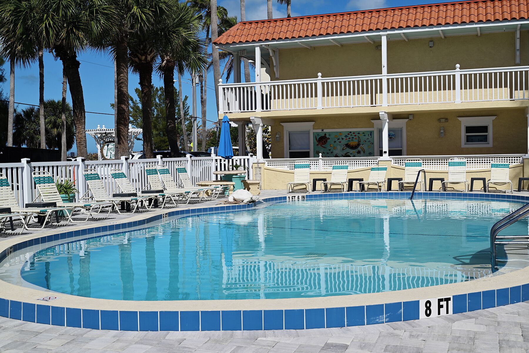A swimming pool with a 3 ft 6 in depth and 8 ft width, surrounded by lounge chairs and a building with a red-tiled roof.