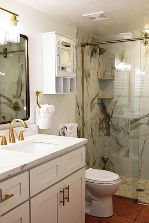 A bathroom with a white vanity, gold fixtures, a glass shower enclosure, and marble walls.