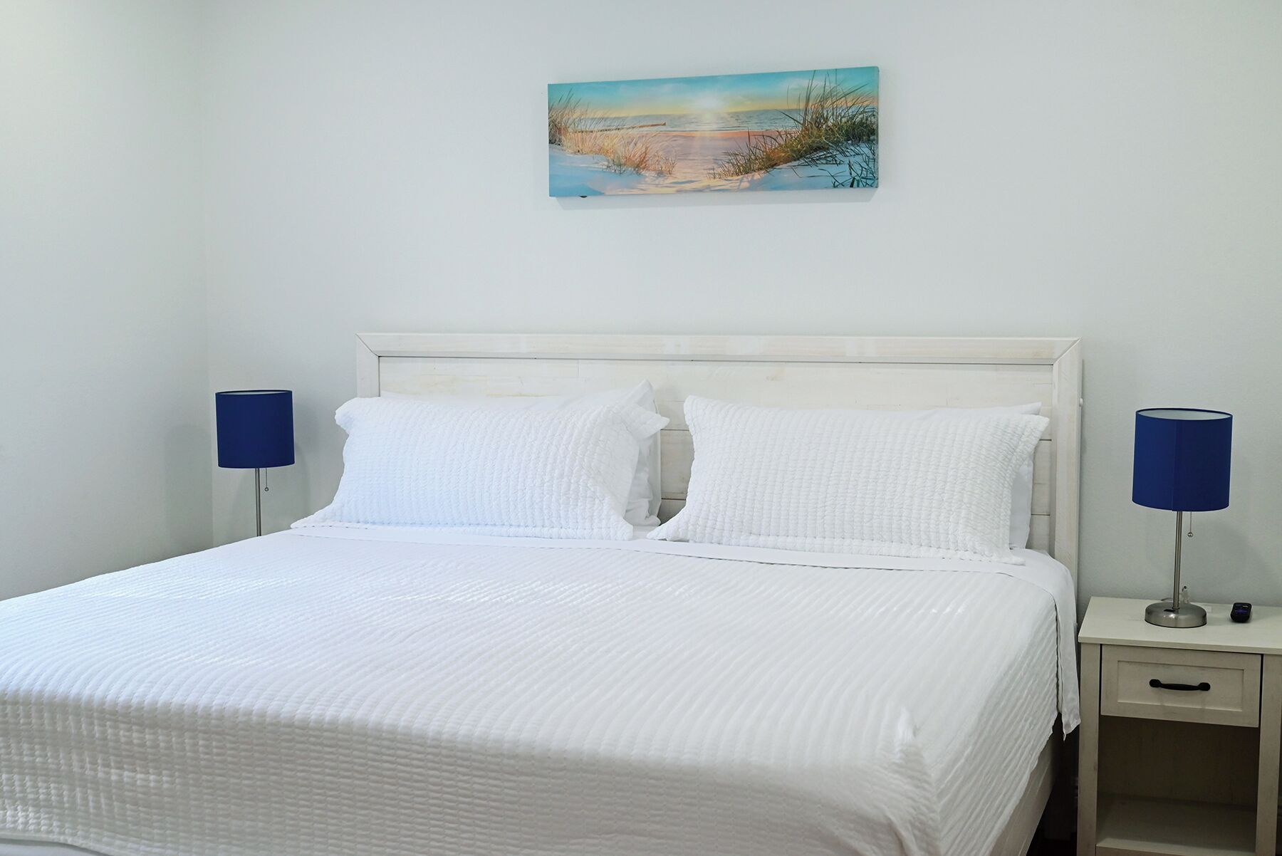 A neatly made bed with white bedding and pillows, flanked by two bedside tables with lamps, in a room with a plain wall and a single piece of wall art.