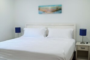A neatly made bed with white bedding and pillows, flanked by two bedside tables with lamps, in a room with a plain wall and a single piece of wall art.