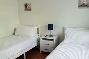 Un dormitorio con dos camas individuales, una mesita de noche con lámpara y un estante pequeño con una planta.