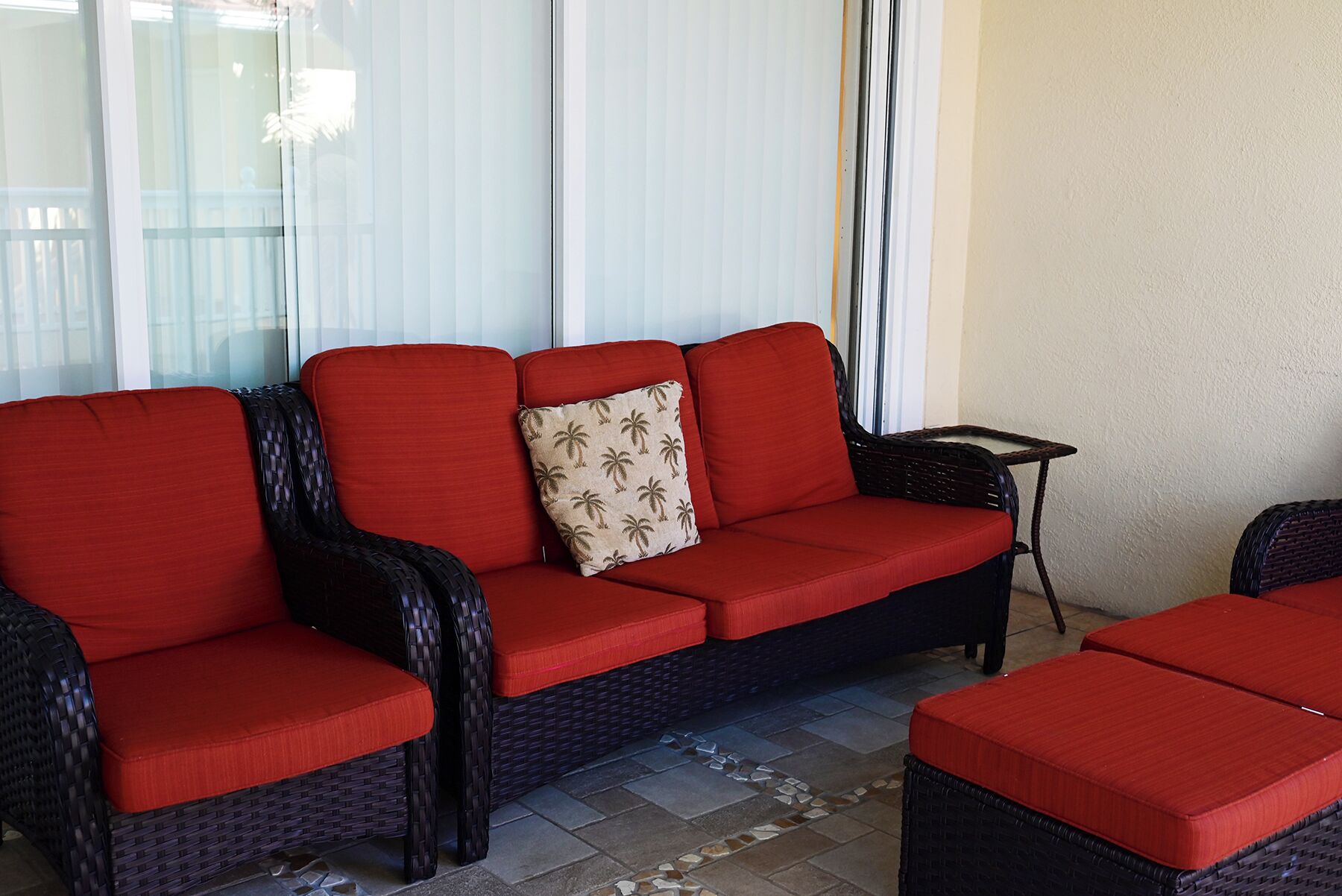 Un sofá rojo con un cojín con forma de palmera, una mesita y sillas de mimbre en un patio empedrado.