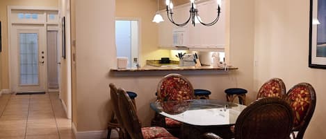 A dining area with a round table, six chairs, a chandelier, and a kitchen in the background.