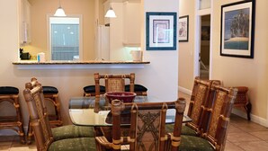 Un comedor con mesa de vidrio, sillas de madera con asientos acolchados, una cocina al fondo y obras de arte enmarcadas en las paredes.