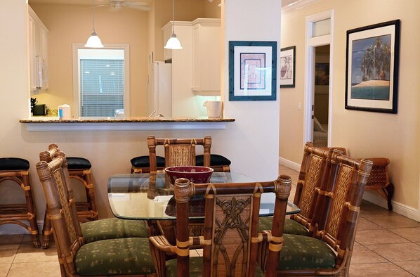 A dining area with a glass table, wooden chairs with cushioned seats, a kitchen in the background, and framed artwork on the walls.