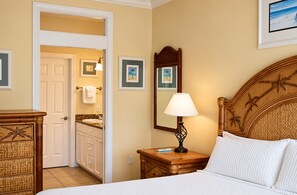 A hotel room with a bed, a nightstand with a lamp, a wooden headboard with palm tree carvings, a bathroom visible through an open door, and framed pictures on the wall.