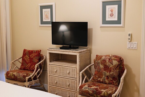 A room with a television on a stand, two wicker chairs with floral upholstery, and framed artwork on the wall.
