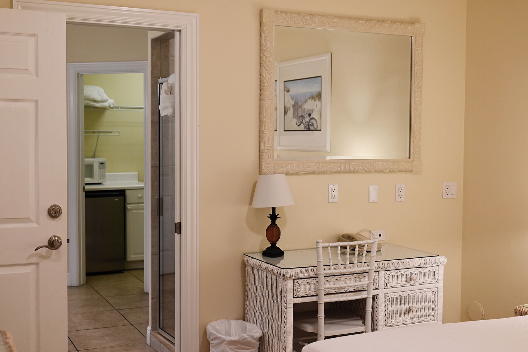 Habitación de hotel con tocador de mimbre blanco, espejo encima y puerta que da a un baño con ducha e inodoro.