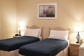 A hotel room with two single beds, a nightstand with a lamp, a framed picture on the wall, and a small wicker side table.