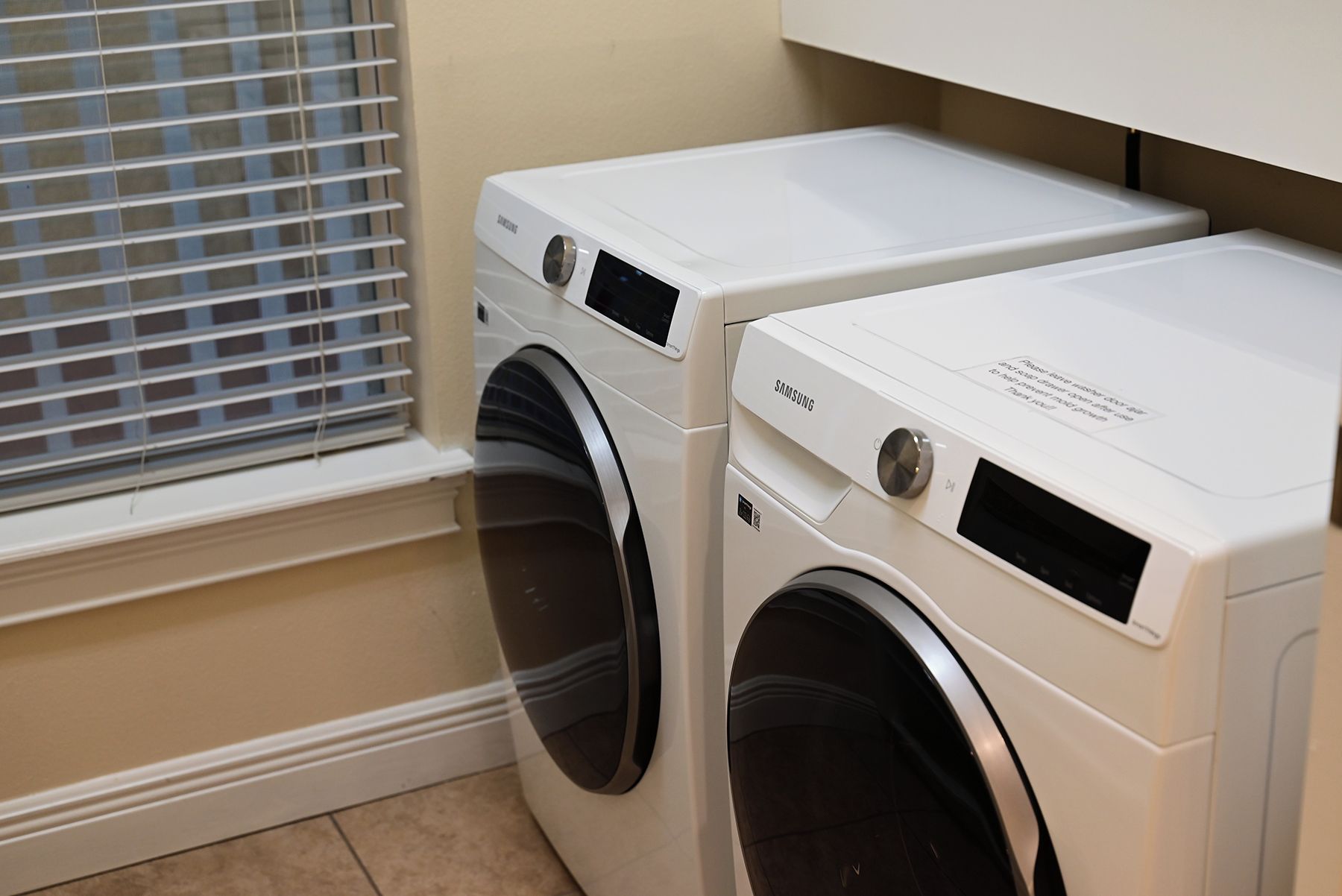 Two white front-loading washing machines in a laundry room with a window and a cabinet above.