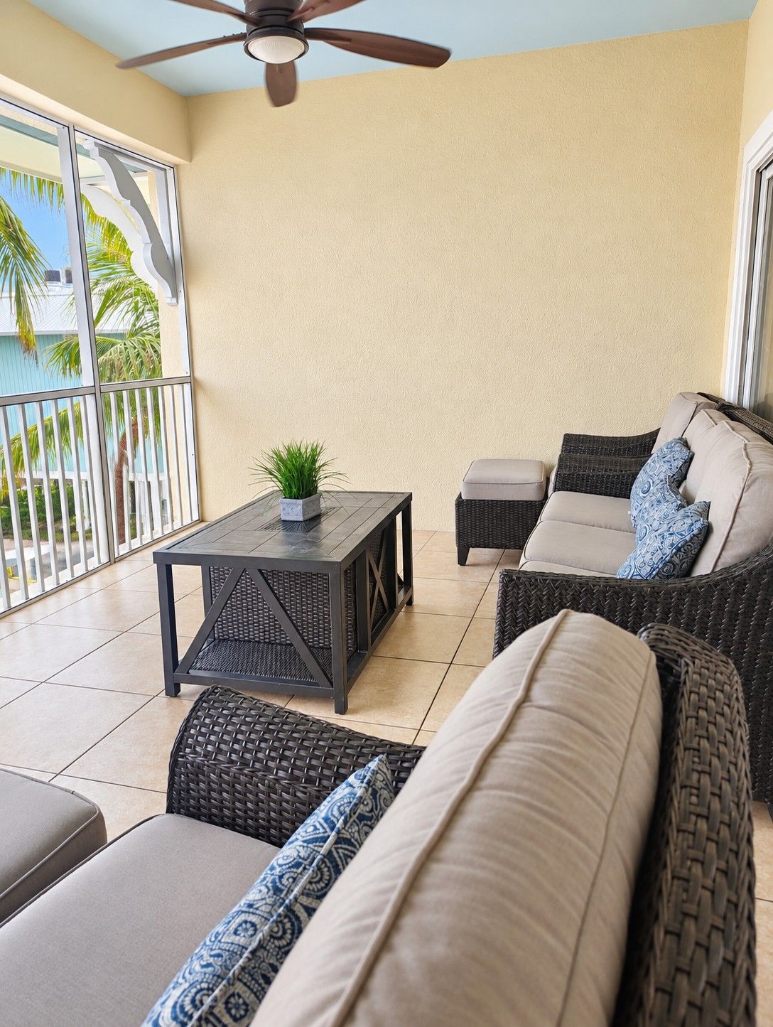 A sunlit patio with wicker furniture, a ceiling fan, and a view of palm trees.