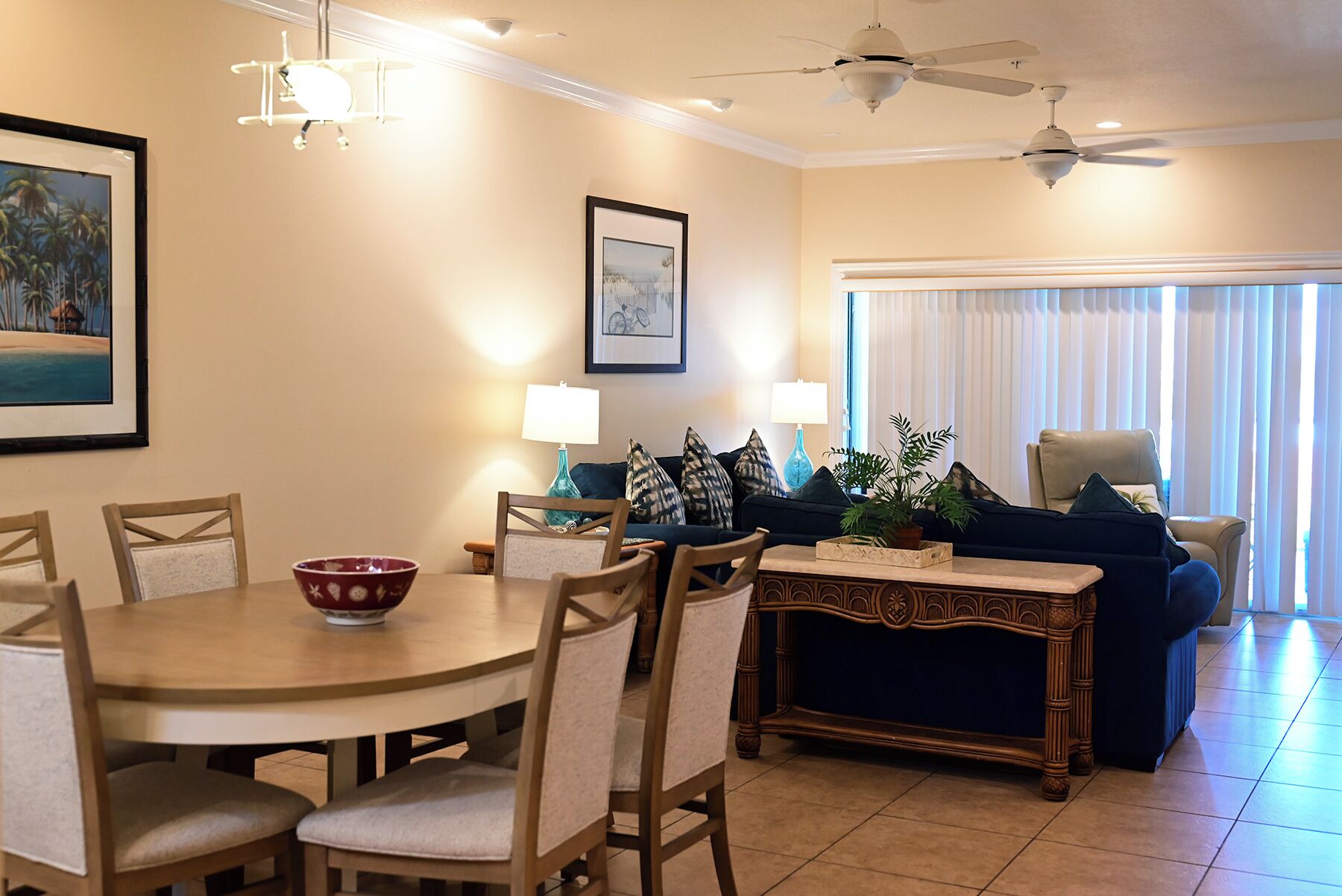 A dining area with a round table, chairs, and a decorative console table. There are ceiling fans, wall-mounted lamps, and a large window with blinds.
