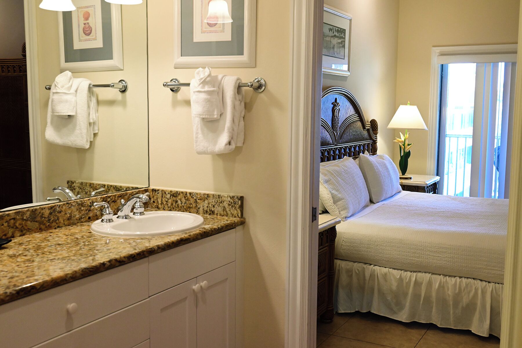 A hotel room with a bathroom, a bed with white bedding, a nightstand with a lamp, and a window with curtains.