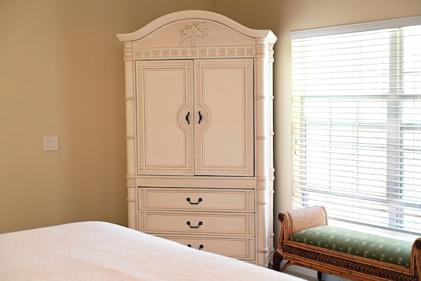 A bedroom with a large white wardrobe, a single bed, a wicker chair with a green cushion, and a window with blinds.