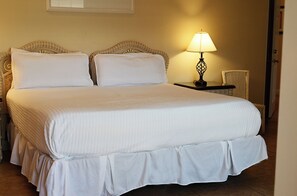 A double bed with a white bedspread and pillows, a bedside table with a lamp, and a chair in a hotel room.