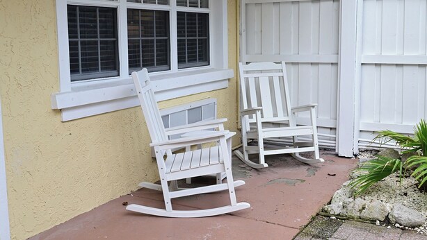 Deux fauteuils à bascule blancs sont placés à l’extérieur d’un bâtiment dont le mur est jaune et qui est entouré d’une clôture blanche.