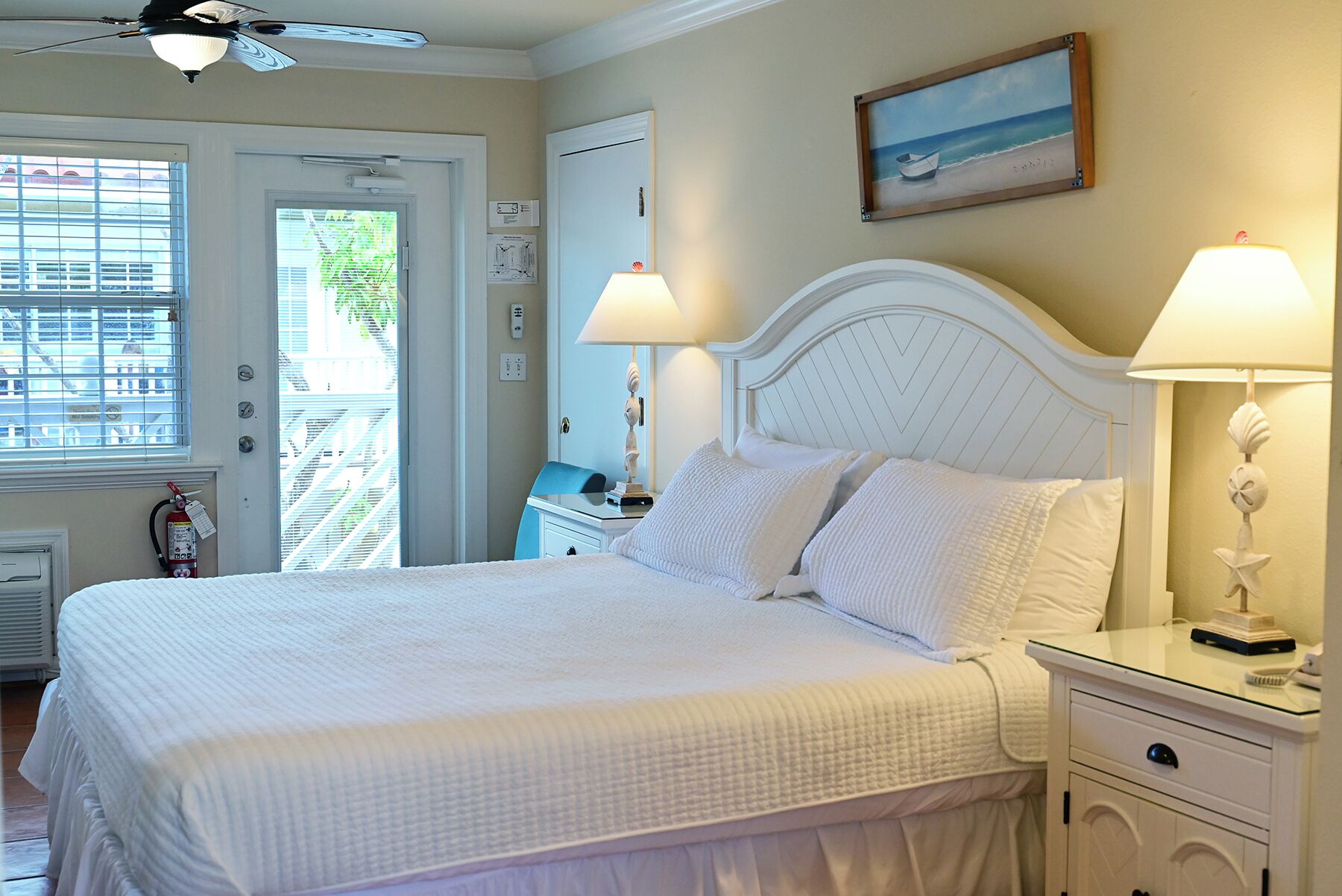 A hotel room with a bed, bedside lamps, a nightstand, a window with blinds, and a painting on the wall.