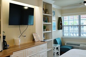 Habitación de hotel con televisión de pantalla plana en la pared, cafetera en la encimera, una pequeña mesa con una silla y una ventana con persianas.