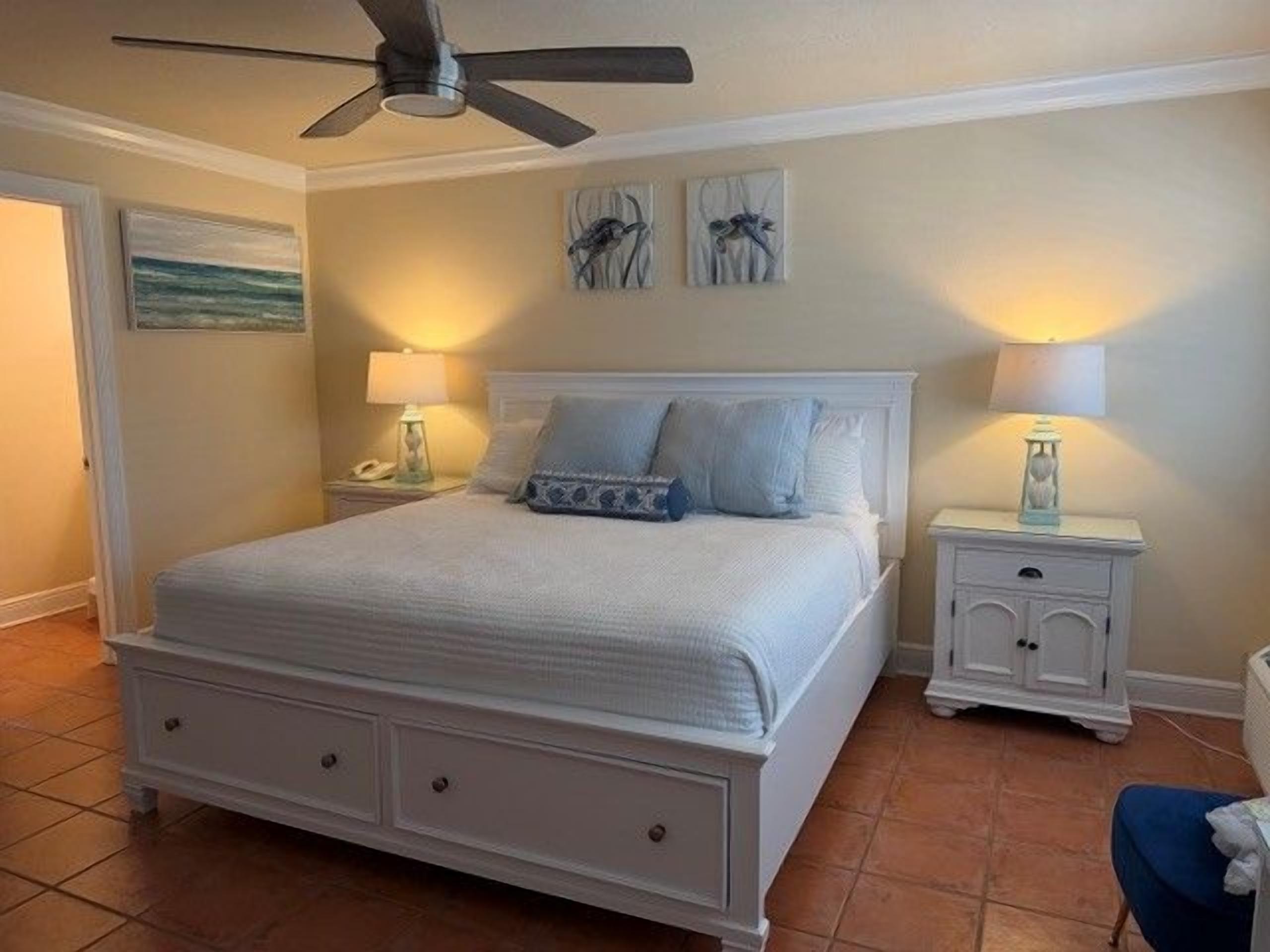 A bedroom with a bed, nightstands, a ceiling fan, and artwork on the walls.