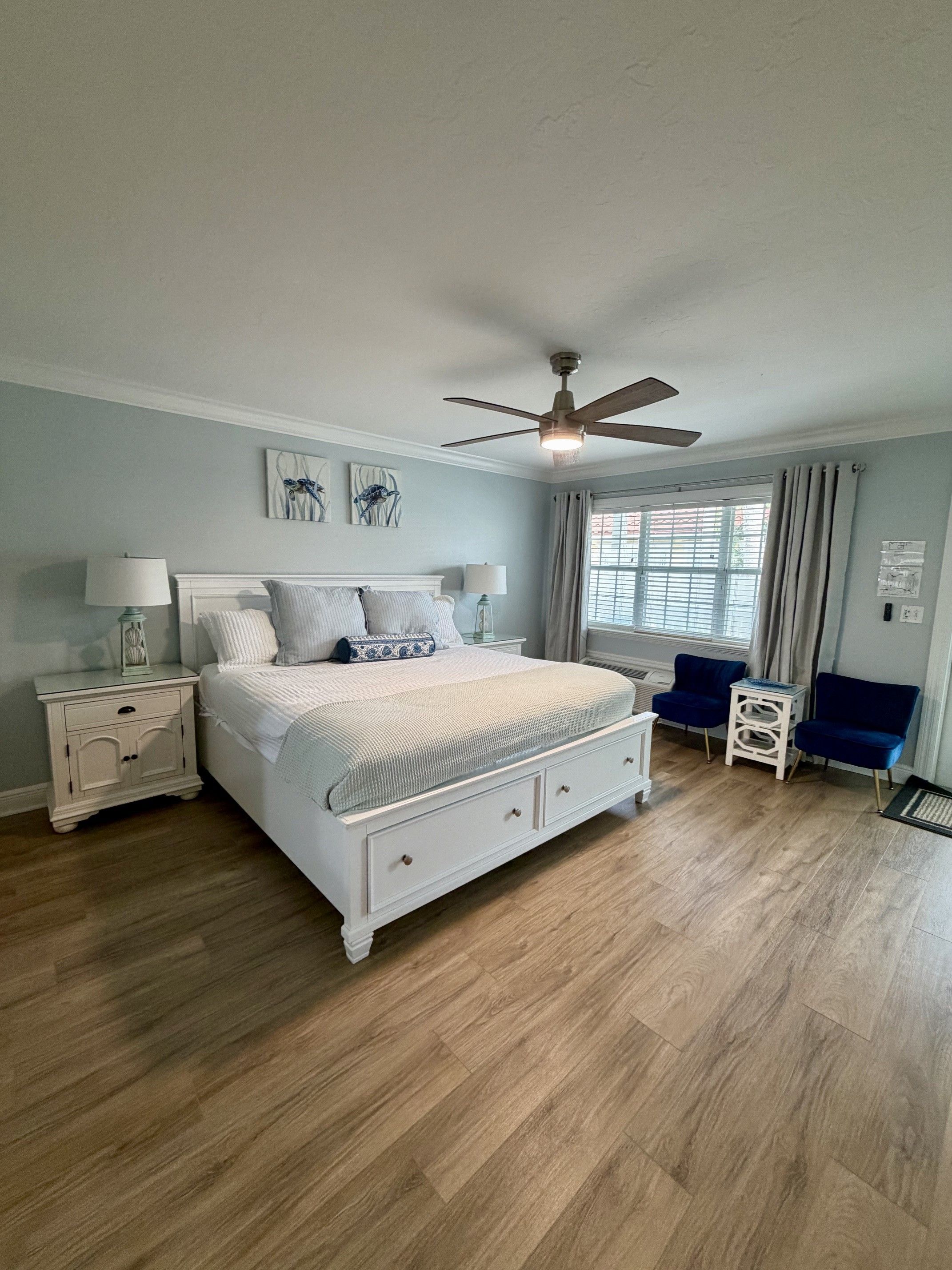 A bedroom with a large bed, a ceiling fan, a window with curtains, and a nightstand with a lamp.