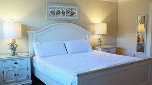 Un lit bien fait, avec une literie blanche, deux tables de chevet avec des lampes, un miroir fixé au mur et un tableau encadré au-dessus du lit.
