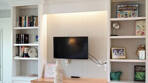 Une pièce comprenant un téléviseur fixé au mur, un bureau en bois et des étagères remplies de livres et d’objets décoratifs.