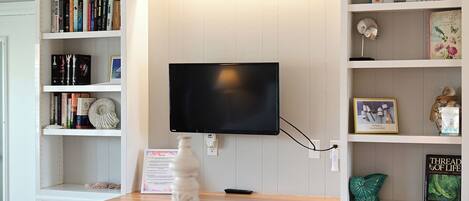A room with a mounted television, a wooden desk, and shelves with books and decorative items.