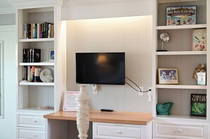 Une pièce comprenant un téléviseur fixé au mur, un bureau en bois et des étagères remplies de livres et d’objets décoratifs.
