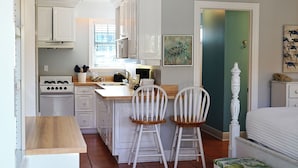 Een compacte keuken met witte kastjes, een fornuis en een spoelbak. Er staan twee witte stoelen bij een kleine tafel. Op de achtergrond is een groene deur te zien.