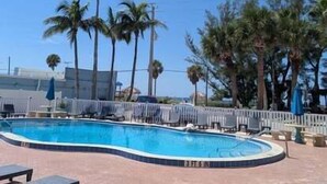 Una piscina de borde curvo, rodeada de palmeras y una valla blanca.