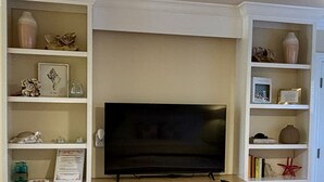 Una habitación con una televisión sobre un soporte de madera, estanterías empotradas blancas con objetos decorativos y muebles blancos con cajones.