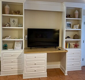Una habitación con una televisión sobre un soporte de madera, estanterías empotradas blancas con objetos decorativos y muebles blancos con cajones.