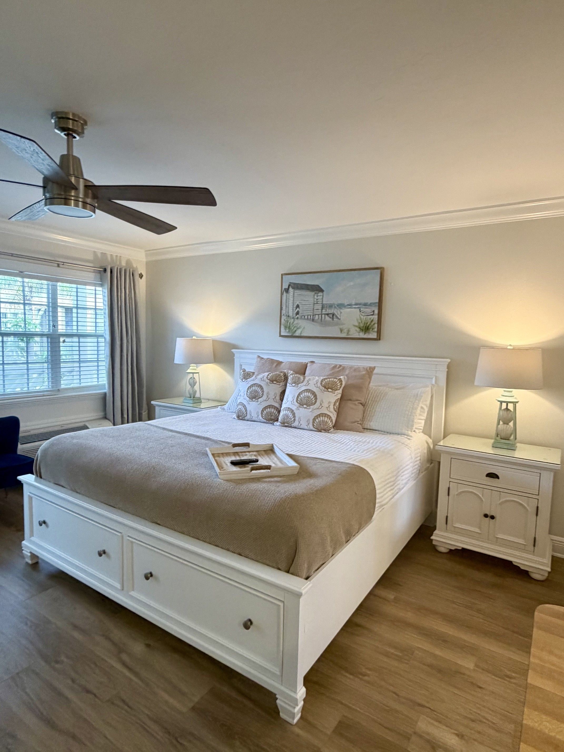 Une chambre à coucher comprenant un lit, deux tables de chevet, un ventilateur de plafond, une fenêtre avec des rideaux et un tableau accroché au mur.