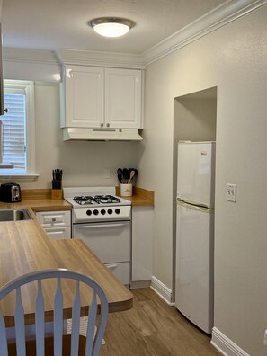 A compact kitchen with white cabinets, a gas stove, a refrigerator, and a wooden dining table with a chair. - 248A: Anna Maria Island, King Studio, Steps from the Beach. (Bradenton Beach)