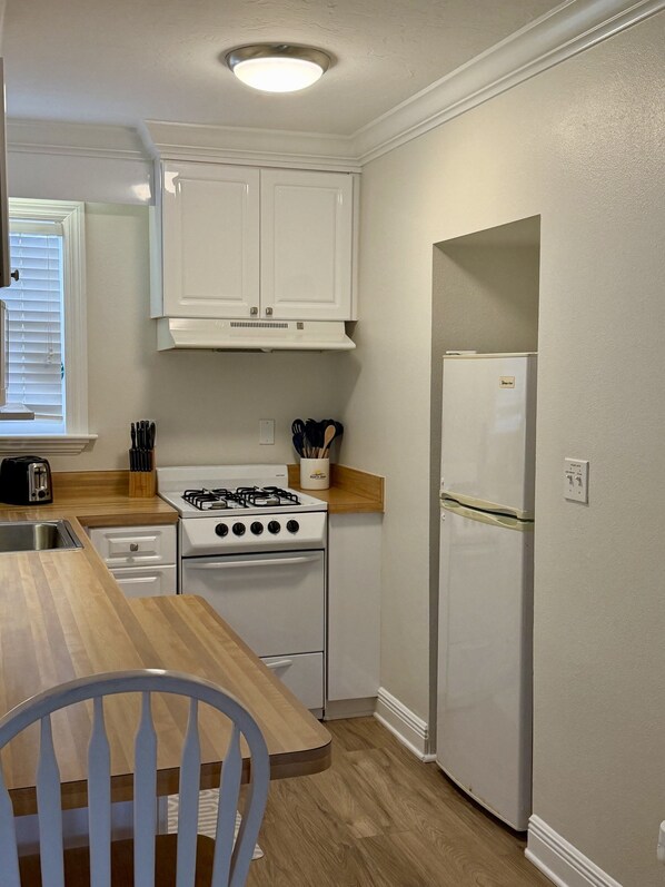 Une cuisine compacte avec des meubles blancs, une cuisinière à gaz, un réfrigérateur et une table à manger en bois avec une chaise.
