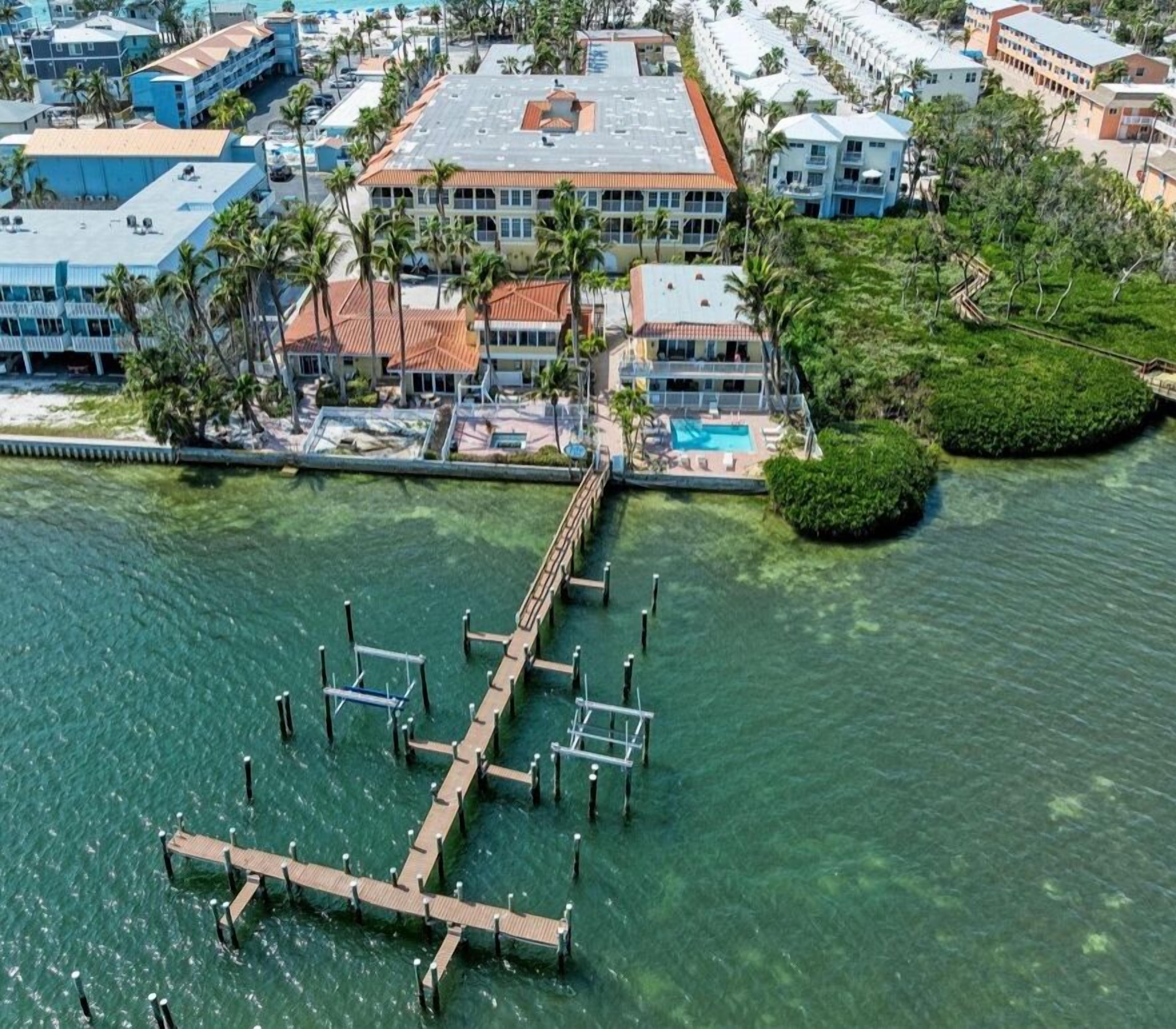 An aerial view of a coastal hotel with a dock extending into the water, surrounded by buildings and palm trees.