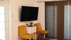 A room with a flat-screen TV mounted on the wall, two mustard-colored chairs, a small white side table with drawers, and a sliding glass door with a brown curtain.