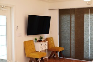 Habitación con televisión de pantalla plana montada en la pared, dos sillas color mostaza, una mesita auxiliar blanca con cajones y una puerta corredera de vidrio con cortina marrón.