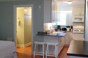 A kitchen with white cabinets, a stove, a microwave, and a sink. There are two white stools and a countertop with various items.